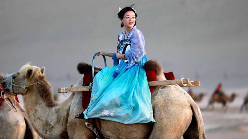 This photo taken on August 10, 2019 shows a woman wearing traditional clothes riding a camel in the desert in Dunhuang in China's northwestern Gansu province. (Photo by STR / AFP) / China OUT