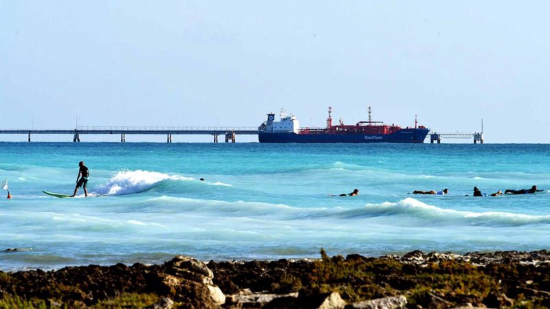 A man surfs as a ship sails next to the "Spiagge Bianche" (white sand) beach, in Rosignano Solvay, a town in Italy's central Tuscany region, on July 31, 2019. - The white sand and unusual Caribbean appearance is not natural, but due to production waste from the Solvay chemical plant in Rosignano Solvay, which produces basic chemical products such as sodium carbonate, bicarbonate, hydrogen peroxide, calcium chloride and chlorine.  AFP/RSS.