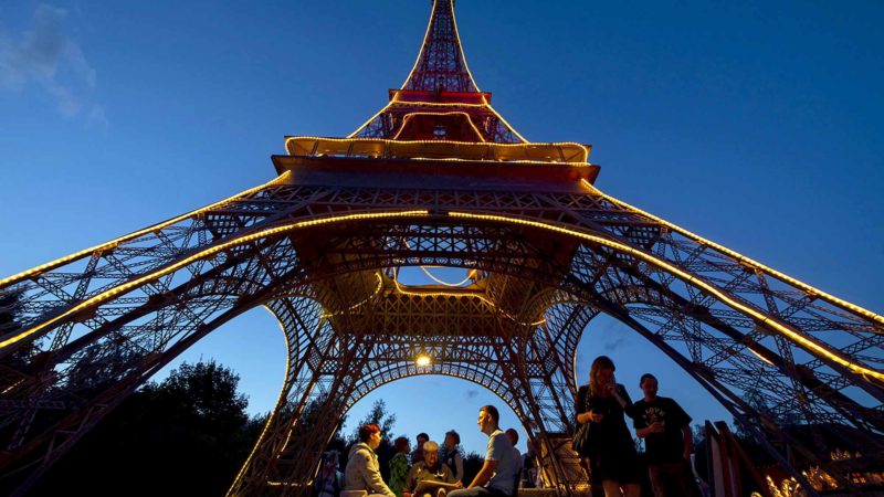 In this picture taken with a long time exposure people walk under an illuminated model of the Eiffel Tower of Paris (12 meters high) in the landscape park Miniwelt (Miniworld) at the event "Miniworld at Night" in Lichtenstein, Germany, Saturday, Aug. 10, 2019. The cultural park Miniworld presents about 100 original and true-to detail buildings and technical facilities at a 1:25 scale ranging on an area of 6,5 hectare. (AP Photo/Jens Meyer)