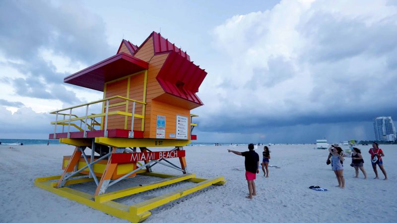 Beach goers on South Beach check out one of the iconic lifeguard stands as bands of rain fall in the distance, Friday, Aug. 30, 2019, on Miami Beach, Fla. All of Florida is under a state of emergency and authorities are urging residents to stockpile a week's worth of food and supplies as Hurricane Dorian gathers strength and aims to slam the state as soon as Monday as a Category 4 storm. (AP Photo/Wilfredo Lee)