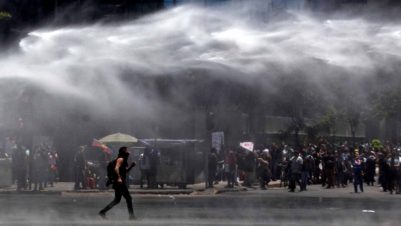 TOPSHOT - A demonstrator is seen in the street as riot police sprays water to disperse the crowd during a protest against the Chilean government in front of La Moneda presidential palace in Santiago on November 26, 2019. - Furious Chileans have since October 18 been protesting social and economic inequality, and against an entrenched political elite that comes from a small number of the wealthiest families in the country, among other issues. (Photo by CLAUDIO REYES / AFP)