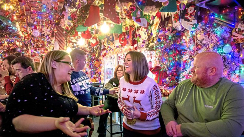 Friends follow a holiday tradition by gathering under approximately 10,000 lights decorating Bob's Garage in O'Hara Township, Pa., Tuesday, Dec. 24, 2019. From left are Erica Lelis, of Springdale, Pa., Katie McGregor, of Ingram, Pa., and Jack Ingraham, of Stowe Township, Pa. The three are part of a group that, for nearly two decades, has gathered at the bar each year on the day before Christmas. The small bar is famous for its light display, which takes two weeks to install. (Steve Mellon/Pittsburgh Post-Gazette via AP)
