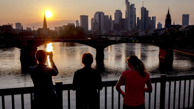 Three women take a look at the buildings of the banking districts at the river Rhine as the sun sets, in Frankfurt, Germany, Thursday, March 19, 2020. Heavy economical losses are expected due to the coronoavirus. Only for most people, the new coronavirus causes only mild or moderate symptoms, such as fever and cough. For some, especially older adults and people with existing health problems, it can cause more severe illness, including pneumonia. (AP Photo/Michael Probst)