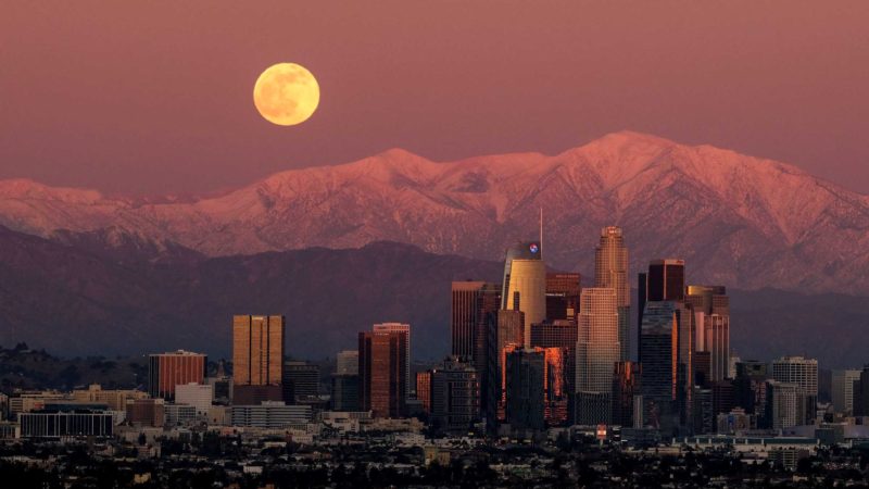 The moon rises over snow-covered mountains, behind the downtown Los Angeles skyline as seen from Kenneth Hahn State Recreation Area Tuesday, Dec. 29, 2020, in Los Angeles. (AP Photo/Ringo H.W. Chiu)