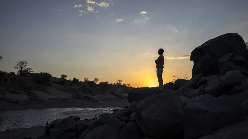 Surgeon and doctor-turned-refugee, Dr. Tewodros Tefera, stands at the Tezeke River crossing in Hamdayet, Sudan, on March 16, 2021. At left across the river is Ethiopia. He plans to continue his work in Sudan. The high-rise buildings of his city in Tigray, Humera, can be seen on the horizon. He could walk home, but he’s not sure he will ever go there again. (AP Photo/Nariman El-Mofty)