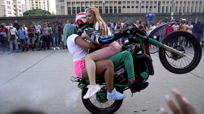 A motorcyclist, with two passengers, performs a wheelie on his motorbike during an exhibition in the El Valle neighborhood of Caracas, Venezuela, Saturday, July 31, 2021. (AP Photo/Ariana Cubillos)