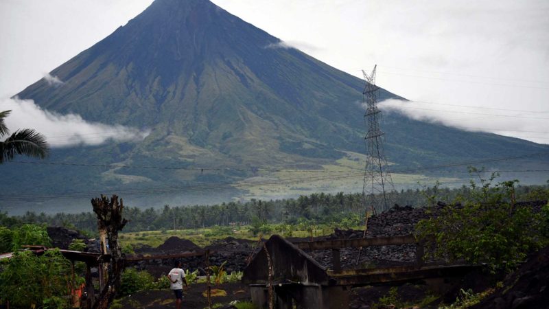 This photo taken on October 5, 2021 shows a resident (L) standing next to a house half-buried by displaced sand and rock brought from the Mayon volcano (background) after heavy rains from Typhoon Goni in 2020 in San Francisco village in Guinobatan, Albay province, south of Manila. - Hundreds of families from poor villages around Mayon volcano in Albay province on the country's most populous island of Luzon are waiting for new homes after Typhoon Goni pounded the region last November. (Photo by Ted ALJIBE / AFP) / TO GO WITH Climate-UN-COP26-Philippines-disaster,PROFILE by Allison Jackson